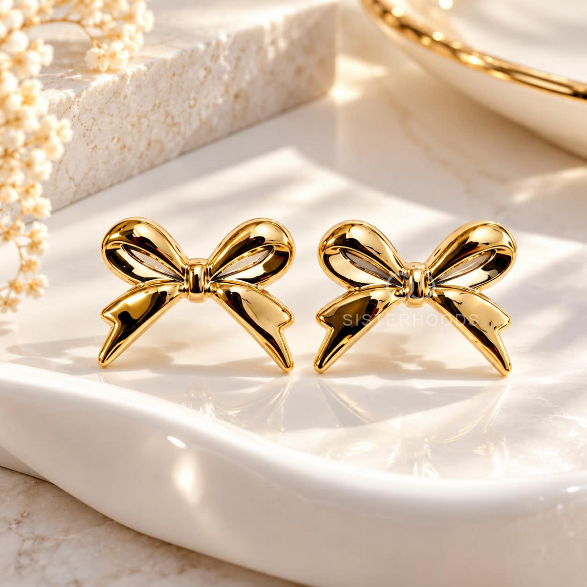 Glossy gold-tone bow stud earrings worn on an ear | SISTERHOODS-SHA256:6b69a35cd56dd208f45236fd678bfde398a7aa6d5ae1d30334327dfd12a1dd57